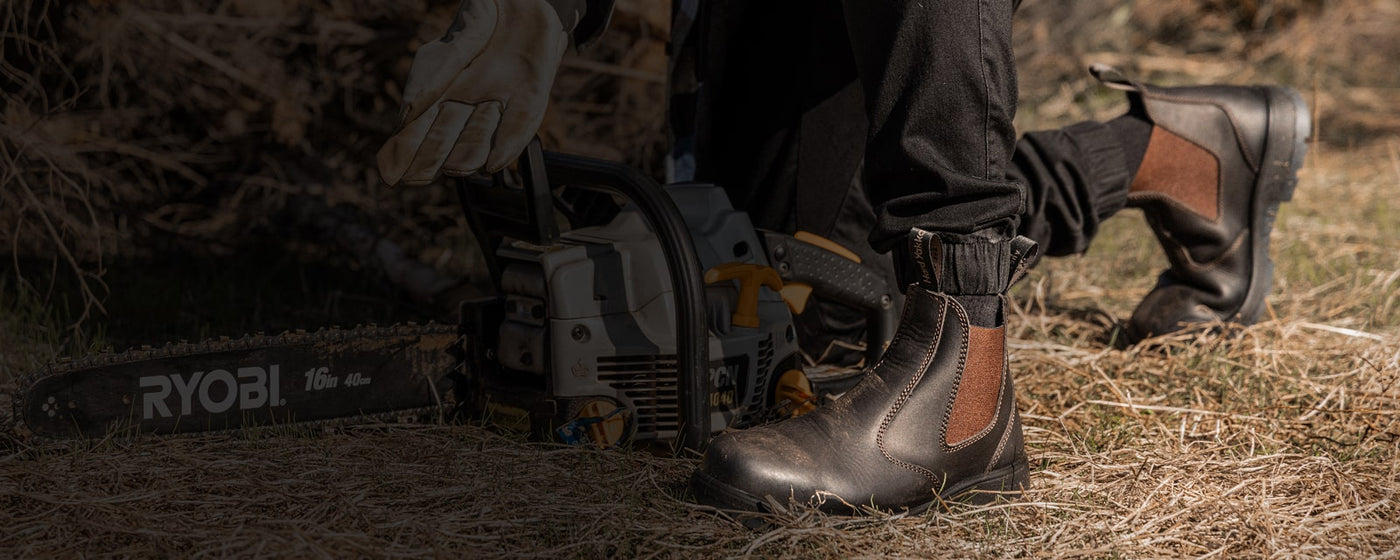 Person kneeling down on hay with a chainsaw, wearing a pair of brown Hard Yakka Outback Brumby dealer boots