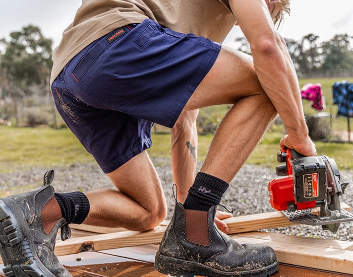 Close-up image of a man kneeling down, sanding wood with a power tool, wearing a pair of Hard Yakka shorts and safety boots