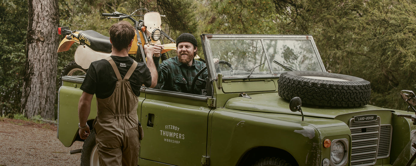 Elton King fist pumping with Brook James, who is driving a land rover with a motorcycle in the back. Both are wearing Hard Yakka workwear